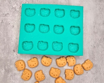 May include: A green silicone mold with 16 cavities shaped like cartoon characters. The mold is on a gray surface with 12 small, brown, cookie-like treats.