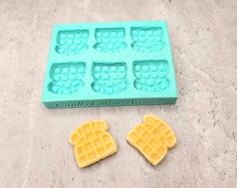 May include: A teal silicone mold with six waffle-shaped cavities. The mold has the text "Vanilla Cotton Aromas" printed on the bottom. Two small, yellow waffle-shaped candies are next to the mold.