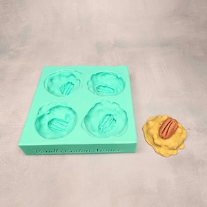 May include: A green silicone mould with four round cavities for making candy. The mould has the text "Vanilla Cotton Armes" on the bottom. A yellow candy with a pecan nut on top is sitting next to the mould.