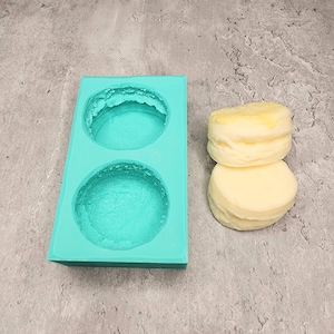 May include: A turquoise silicone mold with two round cavities. Two white and yellow cake rounds are next to the mold.