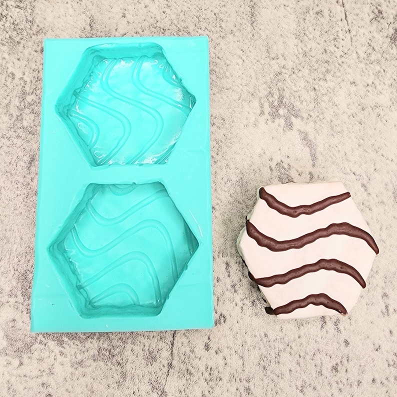 Puede incluir: Un molde de silicona turquesa con dos cavidades hexagonales. El molde est&aacute; dise&ntilde;ado para crear galletas hexagonales u otras golosinas. Una galleta hexagonal blanca con rayas marrones se muestra junto al molde.