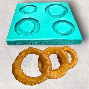 May include: A teal silicone mold with four circular cavities, and the text "Vanilla Cotton Aromas" is above three golden-brown onion rings. The mold is designed for crafting, possibly for soaps or candles.