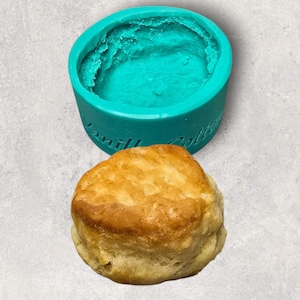 Könnte beinhalten: Ein goldbrauner Keks liegt neben einer türkisfarbenen Silikonform. Die Form ist rund und trägt den Schriftzug "Vanilla Cotton" an der Seite. Der Keks hat eine flockige Textur.