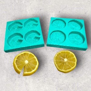 Puede incluir: Dos moldes de silicona turquesa con impresiones de rodajas de limón, junto a dos mitades de limón. Los moldes están etiquetados como "Vanilla Cotton Aromas". Los limones son amarillos con pulpa y semillas visibles, sobre un fondo gris claro.