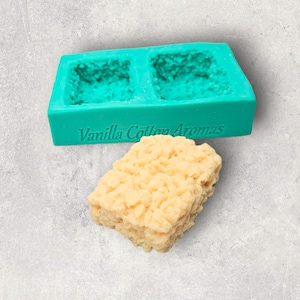 May include: A teal silicone mold with two rectangular cavities. The mold is labeled "Vanilla Cotton Aromas". A light brown, textured bar of soap is sitting next to the mold.