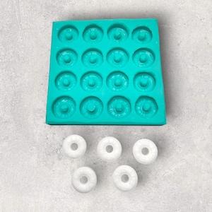 May include: A turquoise silicone mould with 20 round cavities for making doughnuts. Five white doughnuts are shown below the mould.