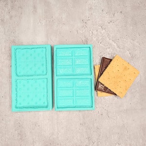 May include: Two turquoise silicone molds for making s'mores. The mold on the left has two square sections for making graham crackers. The mold on the right has two rectangular sections for making chocolate bars. There are two graham crackers and two chocolate bars next to the molds.