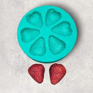 May include: A teal silicone mould with six heart-shaped cavities, designed for making small treats or sweets. Two red strawberries are placed next to the mould.