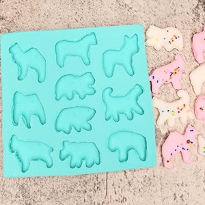 May include: A blue silicone mold with 12 animal-shaped cavities. The mold is filled with colorful, decorated cookies in the shapes of a horse, bear, dog, and other animals.