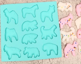 May include: A blue silicone mould with 12 animal-shaped cavities. The mould is filled with colourful, decorated cookies in the shapes of a horse, bear, dog, and other animals.