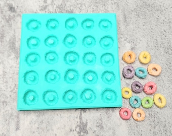 May include: A turquoise silicone mold with 36 round cavities, perfect for making small donuts or other treats.  A variety of colorful cereal rings are scattered on a gray surface.
