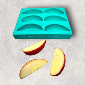 May include: A teal silicone mould with six semi-circular cavities, with the text "Vanilla Cotton Aromas" on the front. Three apple slices with red skin and yellow flesh are below the mould. The image is on a grey background.