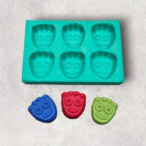 May include: A teal silicone mold with six smiling face cavities, alongside three colorful candies: blue, red, and green. The candies and mold feature a similar design, suggesting a themed treat or craft project.