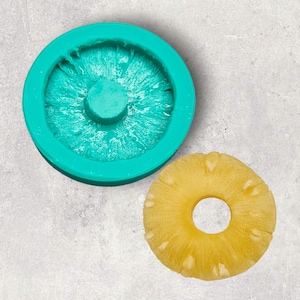 May include: A teal silicone mold shaped like a pineapple slice. The mold is on a gray surface next to a real pineapple slice.