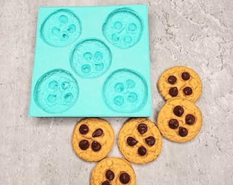 May include: A turquoise silicone mold with six circular cavities, each designed to resemble a cookie. Several baked chocolate chip cookies are arranged around the mold, showcasing the finished product. The cookies have a golden-brown color with dark chocolate chips.