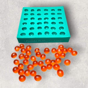 以下が含まれることがあります： 複数の円形のくぼみがある、ターコイズ色のシリコンモールド。ベースには「Vanilla Cotton Aromas」の文字。モールドの周りには、オレンジ色の球状の物体が多数散らばっており、ワックスメルトやバスボムの可能性があります。