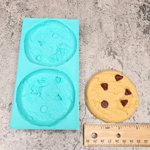 Puede incluir: Un molde de silicona para hacer galletas con chispas de chocolate. El molde es azul claro y tiene dos cavidades con forma de galletas. Una galleta de chocolate real se muestra junto al molde.