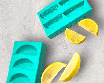 May include: Two turquoise silicone molds for making lemon slices. The molds are rectangular and have three and four sections, respectively. The text "Vanilla Cotton Aromas" is printed on the larger mold. There are also four lemon slices on a gray surface.