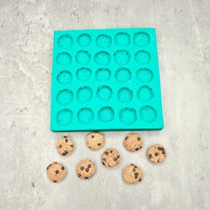Puede incluir: Un molde de silicona turquesa con 36 cavidades redondas, perfecto para hacer pequeñas galletas o caramelos. Hay 9 galletas con chispas de chocolate sobre una superficie gris.