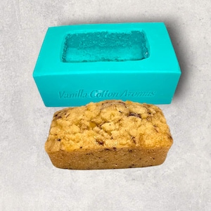 Può includere: Uno stampo per pane in silicone turchese con la scritta "Vanilla Cotton Aromas" sul davanti. Un pane con una copertura di briciole è seduto accanto allo stampo.