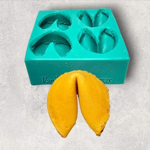 Może przedstawiać: Turkusowa silikonowa forma z czterema odciskami ciasteczek z wróżbą obok złoto-brązowego ciasteczka z wróżbą. Na formie wydrukowano tekst "Van" i "mas". Ciasteczko z wróżbą jest otwarte, odsłaniając wnętrze.