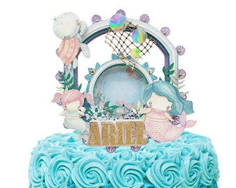 May include: A blue and turquoise ombre cake decorated with buttercream frosting in a rosette pattern. The cake is topped with a mermaid-themed cake topper featuring two mermaids, a net, and the name "Ariel".