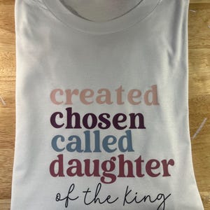 May include: A white t-shirt, folded neatly, featuring the text "created chosen called daughter of the king" in shades of pink, purple, blue, and burgundy. The shirt is made of a soft fabric and has a round neckline.