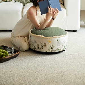 May include: A round green and white floral patterned floor cushion with a fringe trim. The cushion is sitting on a light beige carpet.
