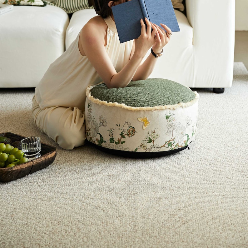 May include: A round green and white floral patterned floor cushion with a fringe trim. The cushion is sitting on a light beige carpet.