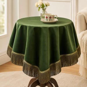 Puede incluir: Un mantel redondo de terciopelo verde oscuro con un borde decorativo de flecos dorados en la parte inferior. El mantel cubre una mesa circular, sobre la cual hay una taza de té blanca con motivos de rosas rojas y un florero de cristal con flores blancas.