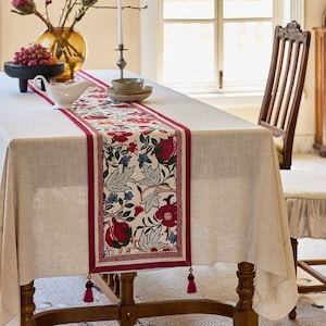 May include: A white tablecloth with a red and white floral table runner with tassels. The table runner features a pomegranate design.