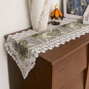 May include: A rectangular table runner featuring a beige fabric center with a repeating pattern of green palm leaves and gray circular motifs. The runner is bordered by an intricate white floral lace trim that hangs over the edges.