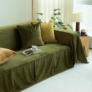 May include: A green velvet sofa with several throw pillows in shades of green and gold. A small teacup and saucer sit on an open book. A lamp and side table are visible in the background. The sofa is covered with a matching velvet slipcover.