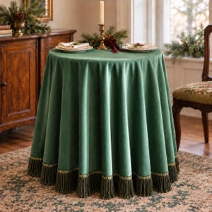 Può includere: Un tavolo rotondo coperto da una tovaglia in velluto verde scuro che arriva fino a terra, rifinita con una frangia decorativa sul bordo inferiore. Il tavolo è apparecchiato con due piatti bianchi, un candelabro in ottone e del verde.