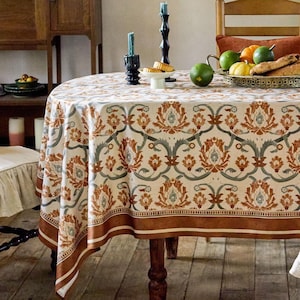 Op de afbeelding: Een ronde tafelkleed met een herhalend bloemmotief in bruin, groen en crèmetinten. Het tafelkleed bedekt een houten tafel, met een bruine rand. De tafel is gedekt met kaarsen, fruit en brood.