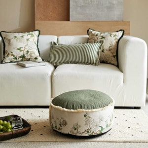 May include: A round green and white floral patterned ottoman with a fringe trim. The ottoman is sitting on a white rug with black dots.