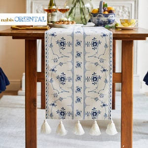 May include: A white table runner with a blue floral pattern and white tassels. The runner is on a wooden table with a bowl of fruit and a glass of wine in the background.