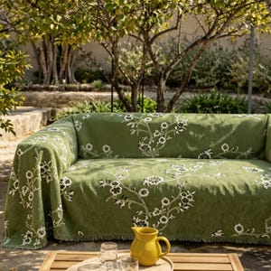 May include: A sage green sofa cover featuring a white floral vine pattern draped over a couch. In the foreground, a yellow ceramic pitcher sits on a wooden table. The fabric has a textured weave and covers the back, seat, and arms of the sofa.