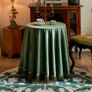 May include: A green velvet table cloth with a gold trim and fringe. The table cloth is on a round table with a cup and saucer and a small decorative box on top.