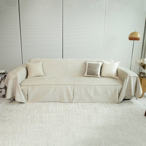 May include: A light beige sofa with a matching throw blanket and three pillows. The sofa has a pleated skirt and is sitting on a light beige rug.