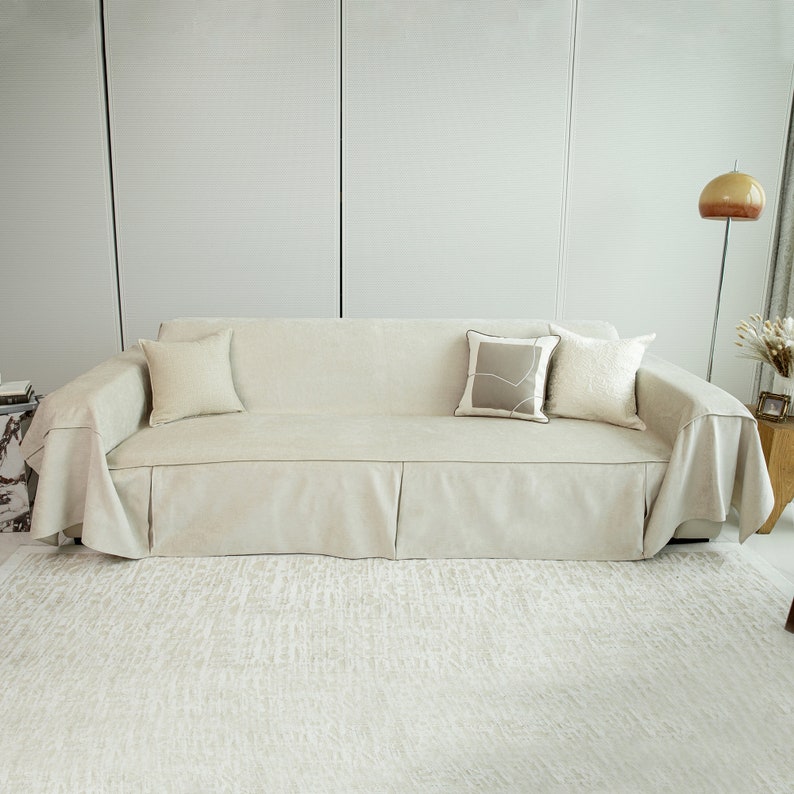May include: A light beige sofa with a matching throw blanket and three pillows. The sofa has a pleated skirt and is sitting on a light beige rug.