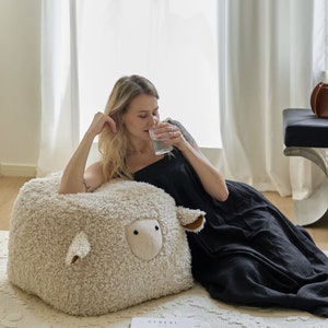 May include: A woman sits on a large, fluffy, white sheep-shaped bean bag chair. She is wearing a black dress and drinking from a clear glass. The bean bag chair is on a white rug.