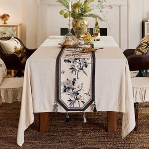 May include: A beige tablecloth with a decorative runner featuring a floral and bird design. The runner has a dark blue border and tassels. The table is set with a centerpiece, glassware, and a covered dish, creating an elegant dining scene.