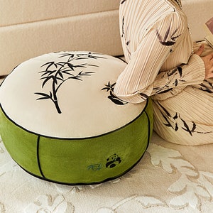 Puede incluir: Un cojín redondo de color crema con un diseño de bambú negro y un borde exterior verde. El cojín tiene un pequeño diseño de panda bordado en el borde verde.