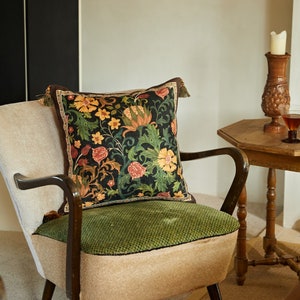 Könnte beinhalten: Ein grüner Samt-Sessel mit einem floralen Kissen. Das Kissen hat einen dunklen Hintergrund mit bunten Blumen und Blättern. Der Sessel steht in einem Raum mit einem Holz-Beistelltisch und einem Kerzenhalter.