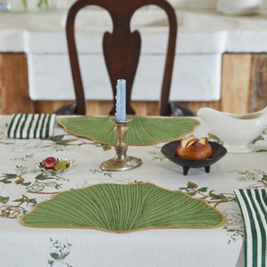 May include: Three green ginkgo leaf-shaped placemats on a white tablecloth with a floral pattern. The placemats have a brown border. A small white pitcher, a brass candlestick with a blue candle, and a small black dish with a brown pastry are on the table.