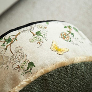 May include: A cream-colored pillow with a floral print and a yellow embroidered butterfly. The pillow has a brown and white fringe around the edge and a dark green fabric backing.
