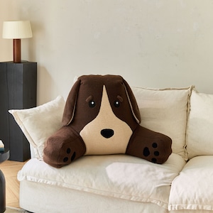 May include: A brown dog-shaped cushion with a white face and black eyes. The cushion is sitting on a white sofa.
