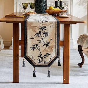 Puede incluir: Camino de mesa rectangular beige con un diseño de bambú negro y borlas decorativas. El camino de mesa está colocado sobre una mesa de madera, con un borde negro y detalles dorados. El diseño de bambú presenta hojas y ramas negras.