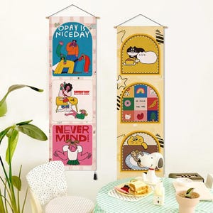 May include: Two vertical fabric wall hangings with wooden rods and black string. The left banner features three panels with cartoon figures and text reading TODAY IS NICEDAY, LOVELY DAY BEAUTIFUL PEOPLE, and NEVER MIND. The right banner shows cats.